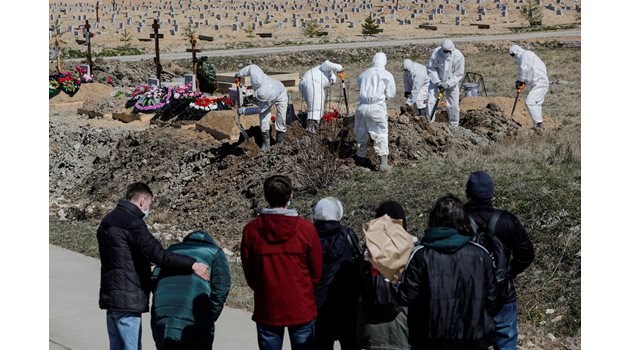 Роднини гледат от далече как гробари в защитни облекла погребват техен близък.