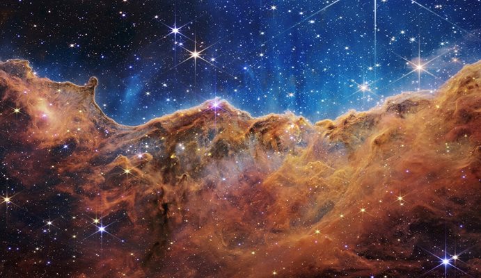 „Космическите скали“ на мъглявината Карина се виждат на изображение, разделено хоризонтално от вълнообразна линия между облачен пейзаж, образуващ мъглявина по протежение на долната част и сравнително ясна горна част, с данни от космическия телескоп Джеймс Уеб на НАСА.
