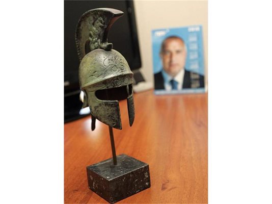 Зад железния древногръцки шлем на бюрото на Радев от снимка строго гледа лидерът Бойко Борисов.