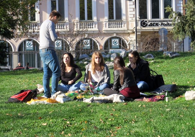 Младежи са насядали на тревата в градинката пред Народния театър в София. В петък температурите в страната бяха между 12 и 19 градуса.